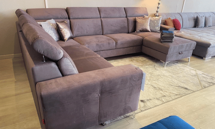 Canapé sectionnel en forme de U, velours mauve/gris-rose, avec repose-pieds et coussins décoratifs, sur plancher clair et tapis texturé.