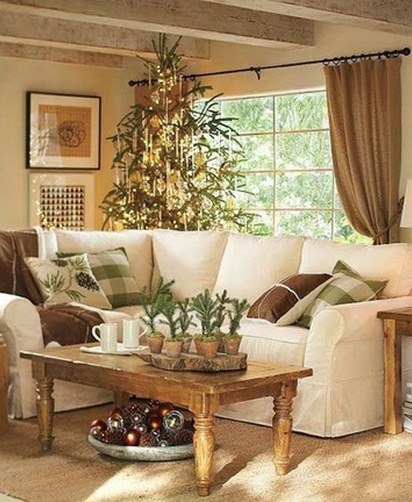 Salon cosy décoré pour Noël avec un grand canapé blanc, sapin éclairé, table basse rustique avec petites plantes et boules décoratives.